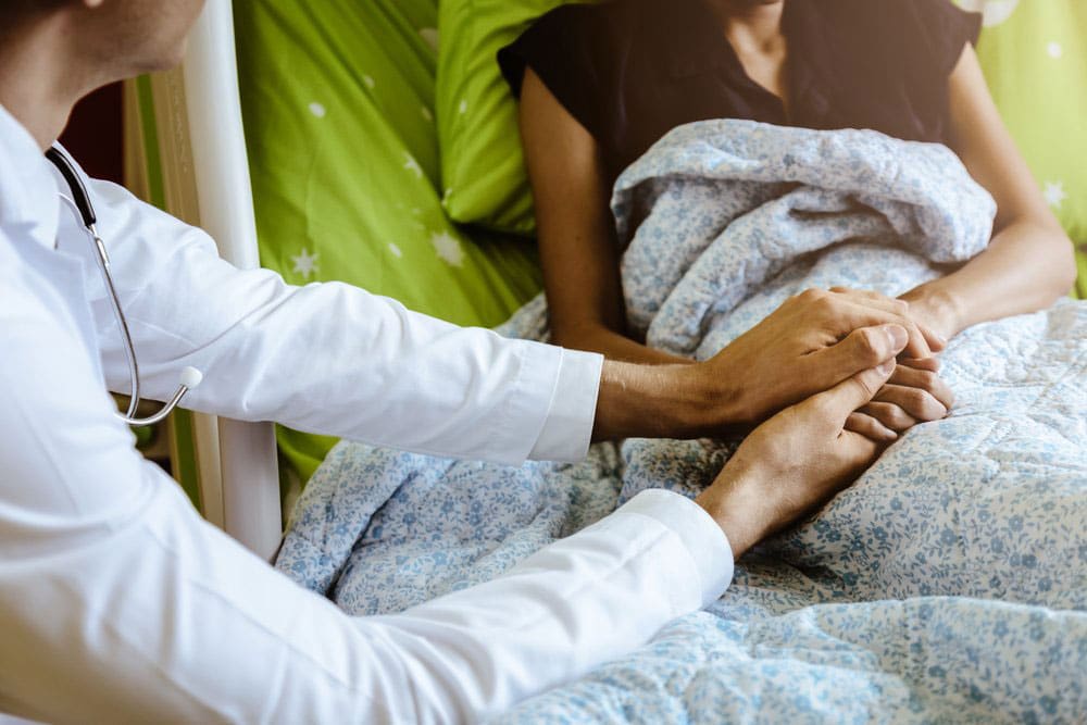 Doctor at bedside of hospice patient at home, holding her hand.