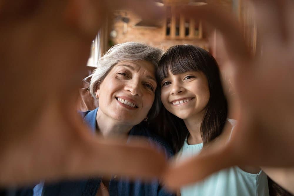 Grandmother and granddaughter smile, with their hands making a heart shape toward the camera.